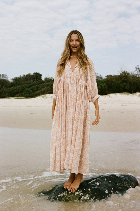 Daughters of India Kyra Maxi Dress in Apricot Blush — standing on rock in surf smiling, sand dunes behind