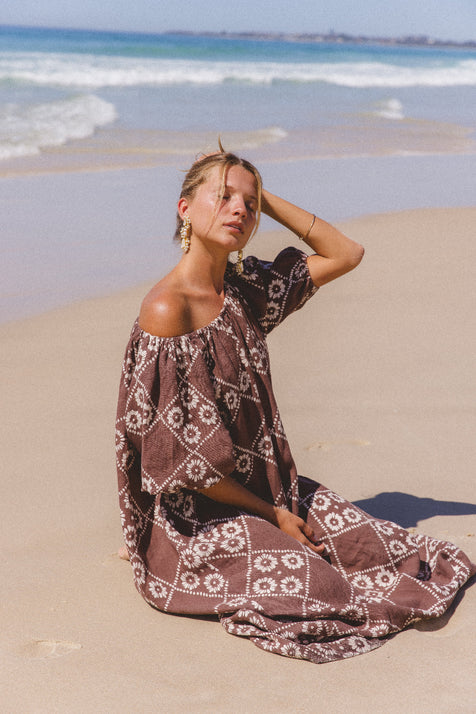 Dahlia Maxi Dress Maple — seated on sandy beach, off-shoulder styling showing hand block print linen, maple geometric floral pattern | Daughters of India
