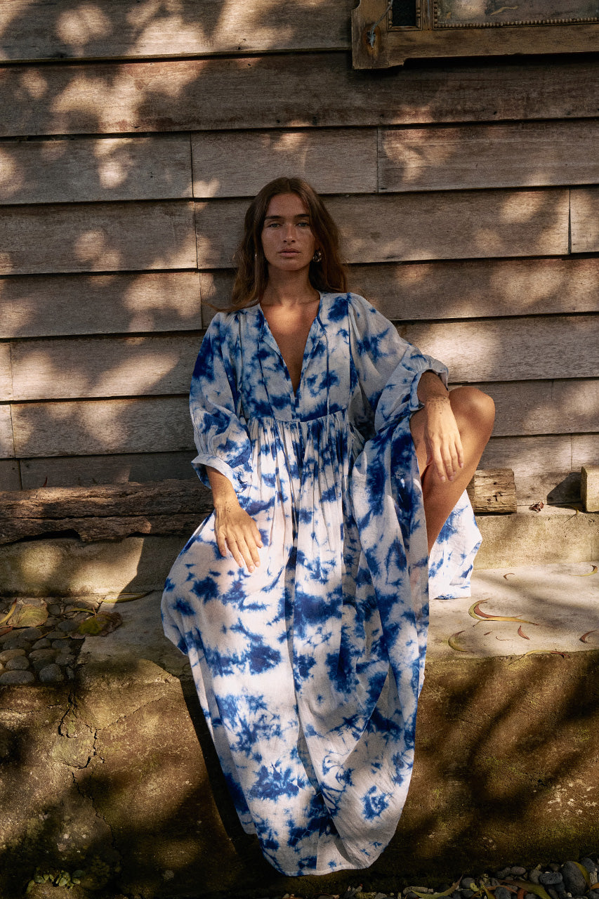 Kyra Maxi Dress Deep Sea — full length seated front view against rustic wooden wall, deep sea shibori tie-dye cotton maxi dress, relaxed flowing silhouette, dappled sunlight | Daughters of India