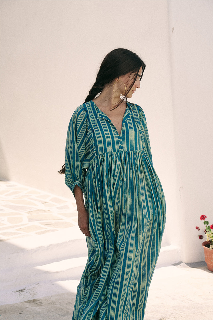 Woman looking downward wearing the Daughters of India Kyra Maxi Dress in Spearmint in a Mediterranean setting, showcasing the relaxed silhouette of the green striped dress